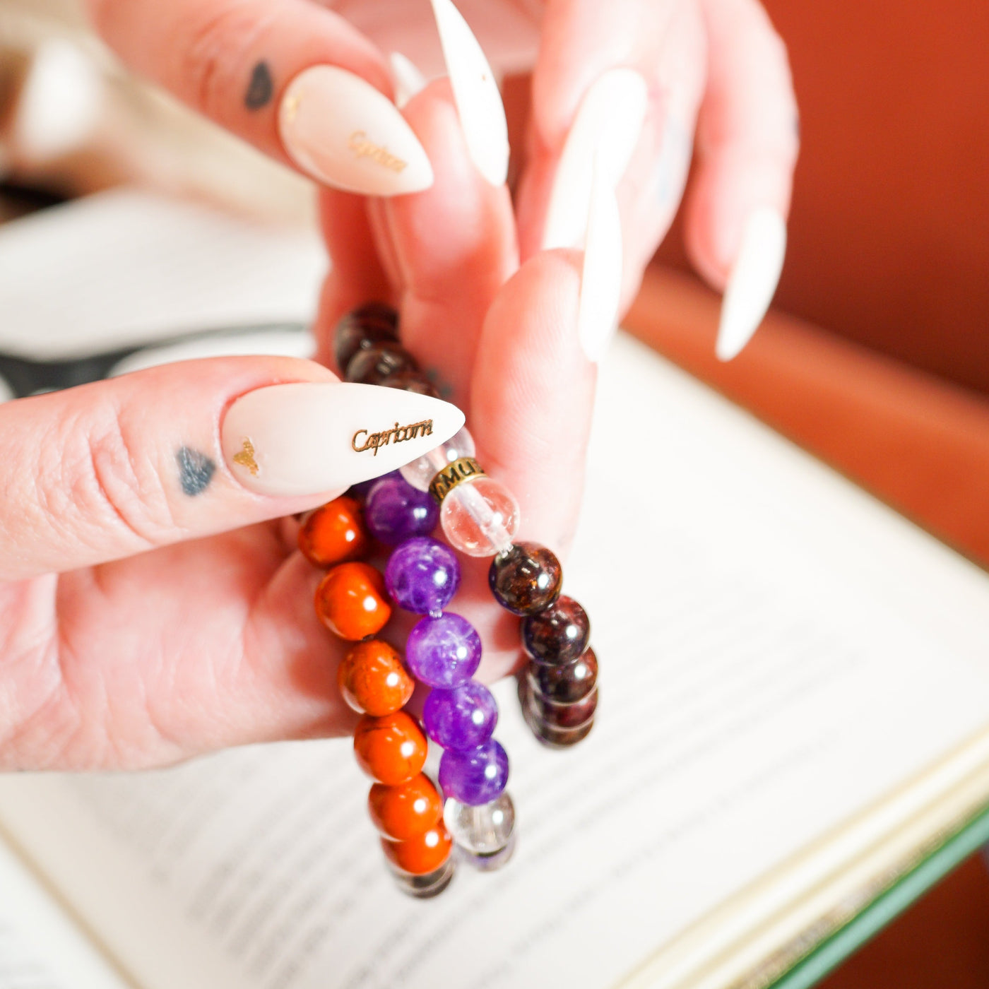 Close up of white woman's hands with pointed white nails that say Capricorn holding an Amethyst, Garnet and Red Jasper bracelets by Energy Muse