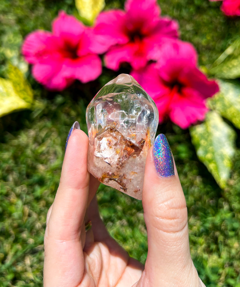 woman holding a semi-clear angel phantom (amphibole) quartz crystal by fuchsia hibiscus flowers and green grass by Energy Muse