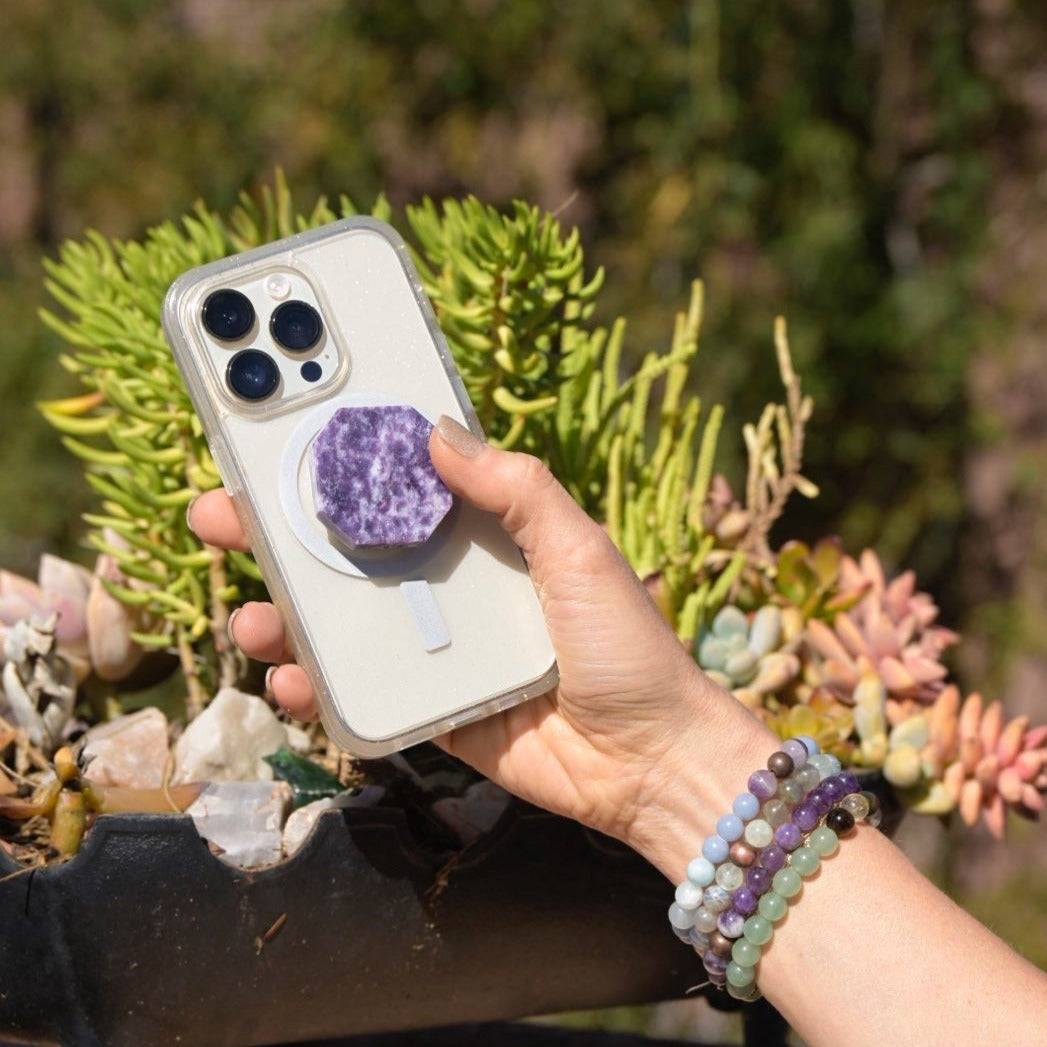 A woman holding her smartphone with Tucson FInd Lepidolite Phone Grip and Stand attached to the phone's case outdoors by Energy Muse