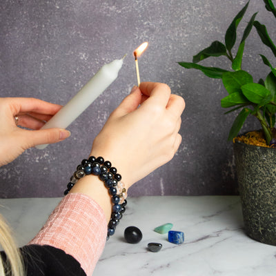 Woman lighting white taper candle in Mercury Retrograde Ritual Kit and wearing Mercury Retrograde bracelet trio by Energy Muse featuring a faceted Sodalite elastic bracelet, a smooth bead Black Tourmaline stretch bracelet and smooth Hematite bead stretch bracelet.