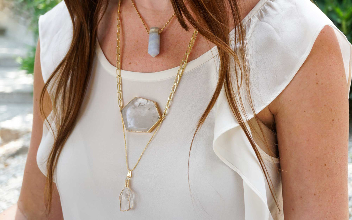 Close up shot of young woman's chest layering a genuine Blue Lace Agate crystal necklace, a wire-wrapped clear quartz crystal necklace, and a large clear quartz necklace on white frilly top by Energy Muse