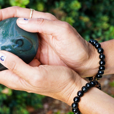A person wearing a Moving Through It bracelet from Energy Muse on each wrist, holding a rainbow obsidian crystal in both hands, outside in nature