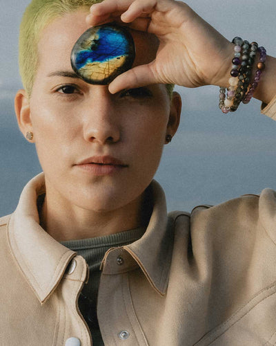 Young non-binary person with short bleached hair holding up genuine Labradorite touchstone to forehead and wearing several Energy Muse crystal bracelets.