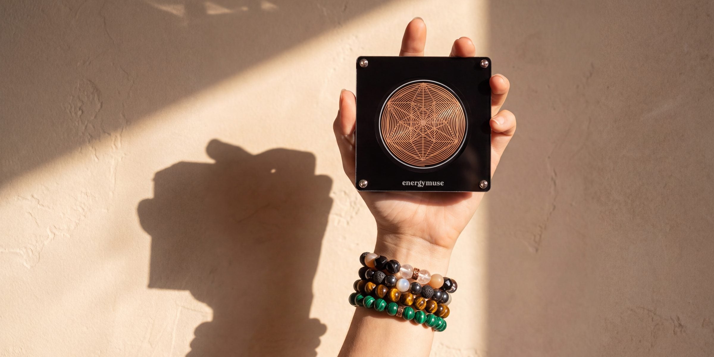 A raised hand holding an Energy Muse frequency generator against a sunlit plaster wall, its shadow falling beside it, crystal bracelets at the wrist